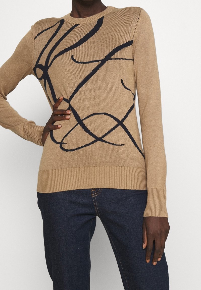 Pull beige à manches longues avec des lignes abstraites noires, associé à un jean bleu foncé, porté par une personne avec la main sur la taille et une posture détendue.
