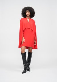 Moteris su garbanotais plaukais, dėvinti raudoną apsiausto suknelę ir juodus aukštus batus iki kelių, stovinti ant šviesaus paprasto fono.