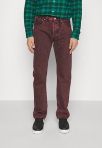 Persona vistiendo jeans rojos con efecto desgastado, zapatillas negras y blancas, y una camisa de cuadros verde, de pie sobre un suelo gris liso.