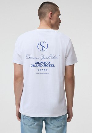Mężczyzna z krótkimi włosami w białej koszulce z niebieskim napisem "Diverse Sport Club MONACO GRAND HOTEL" oraz pięcioma gwiazdkami na plecach.