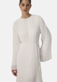 Robe plissée blanche à manches longues et col rond. La taille est marquée par une ceinture assortie, créant une silhouette fluide.