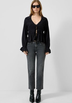 Femme portant un chemisier noir froncé à nouer sur le devant, un jean gris droit, des bottes noires à bout pointu et des lunettes de soleil noires sur fond blanc.