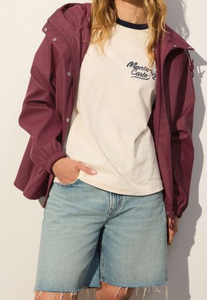 Person wearing a burgundy jacket over a white "Monte Carlo" t-shirt and light blue denim shorts with frayed hems.