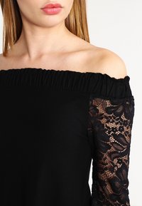 Top nero a spalle scoperte con maniche lunghe in pizzo testurizzato con motivi floreali e scollo arricciato. Tessuto morbido con design attillato.