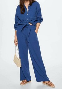 Sininen pellavapaita solmioneurolla ja napituksella, yhdistetty löysälahkeisiin sinisiin housuihin. Asusteina kudottu vaalea laukku ja ruskeat sandaalit.