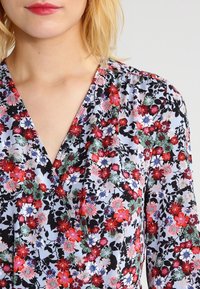 Blusa a stampa floreale con scollatura a V profonda. Presenta piccoli fiori rossi, rosa e blu su uno sfondo azzurro chiaro, con dettagli neri.