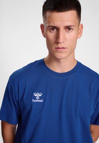 Camiseta de algodón azul con mangas cortas y cuello redondo, que presenta un logo bordado en blanco de "hummel" en el lado izquierdo del pecho.