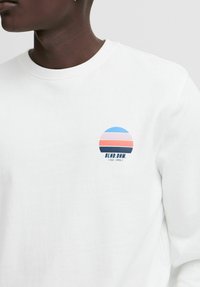 Witte sweatshirt met een rond blauw en roze logo. Heeft een geribde kraag en een casual pasvorm, gemaakt van zacht katoen.