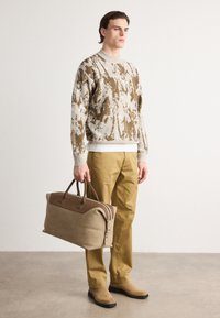 Knit sweater in brown and beige abstract pattern, beige pants, and carrying a tan duffel bag with brown leather accents and handles.