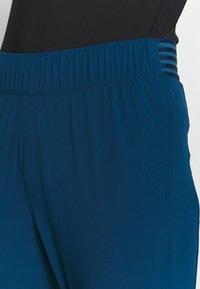 Blå sportbyxor med en bred elastisk midja och mörka randiga detaljer, tillverkade av lätt tyg med en slät textur.