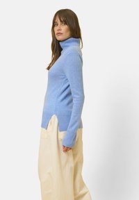 Maglione a collo alto ribbato di colore azzurro chiaro con una vestibilità rilassata, fessure laterali e maniche lunghe, indossato con pantaloni a gamba larga di colore crema.