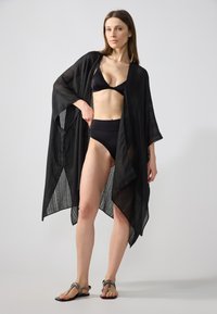 Mustahim mustari hitam dengan bagian depan terbuka dan lengan lebar, menampilkan tekstur bergaris vertikal. Dipakai di atas bikini hitam, dipasangkan dengan sandal datar.