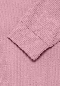 Gros plan sur une manche de pull en maille côtelée rose pliée sur la poitrine montrant la texture et le motif.