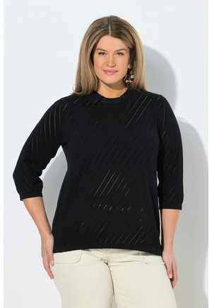 Femme aux cheveux blonds mi-longs portant un haut noir à manches trois quarts et un pantalon crème, posant devant un fond blanc uni.