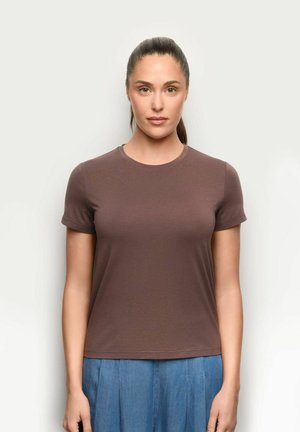 Kobieta z ciemnymi włosami związanymi do tyłu, ubrana w prosty brązowy t-shirt z krótkim rękawem oraz niebieskie spodnie z wysokim stanem, stoi na białym tle.