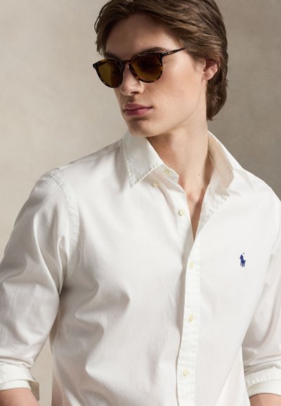 Νέος άνδρας που φοράει γυαλιά ηλίου με μοτίβο καφετί χελώνας και λευκό πουκάμισο με κουμπιά, με ένα μικρό μπλε λογότυπο αλόγου, κοιτάζει ελαφρώς προς τα αριστερά.