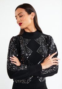 Maglione nero con motivi a diamante in paillettes sulle maniche, colletto alto e polsini a coste, dal texture soffice e dalla vestibilità aderente.