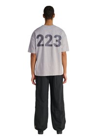 Mand med ryggen til iført grå kortærmet skjorte med stort sort "223" på bagsiden og løse sorte bukser mod hvid baggrund.