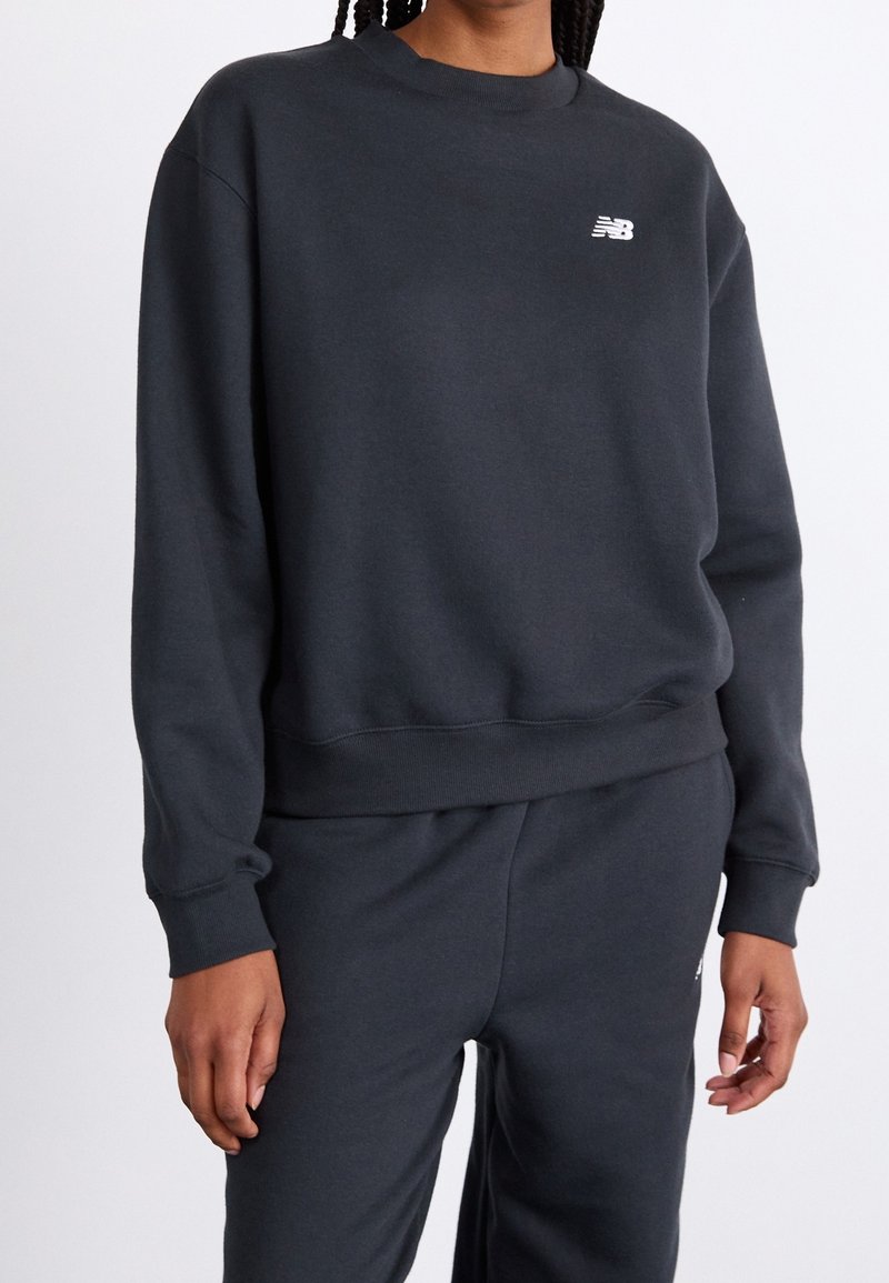 Personne portant un sweat-shirt noir New Balance et un pantalon noir assorti, debout devant un fond blanc uni.