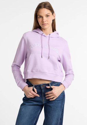 Levanduľová vysoko strihaná mikina s kapucňou, ktorá má biele logo "GUESS JEANS" a prednú klokaní kapsu. Kombinovaná s tmavomodrými džínsami.