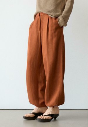 Žena v hnědočervených volných kalhotách, béžovém svetru a černých sandálech na podpatku stojí před jednoduchým světlým pozadím.