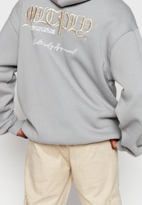 Szara bluza z kapturem z dużą haftowaną grafiką na plecach w białym i złotym kolorze, zawierającą tekst i logo. Połączona z jasnobeżowymi spodniami.