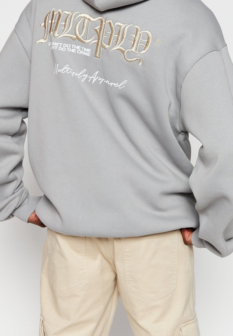 Szara bluza z kapturem z dużą haftowaną grafiką na plecach w białym i złotym kolorze, zawierającą tekst i logo. Połączona z jasnobeżowymi spodniami.