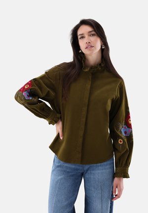 Femme aux longs cheveux foncés portant un chemisier vert olive avec broderies florales sur les manches et un jean bleu, debout avec la main sur la hanche.