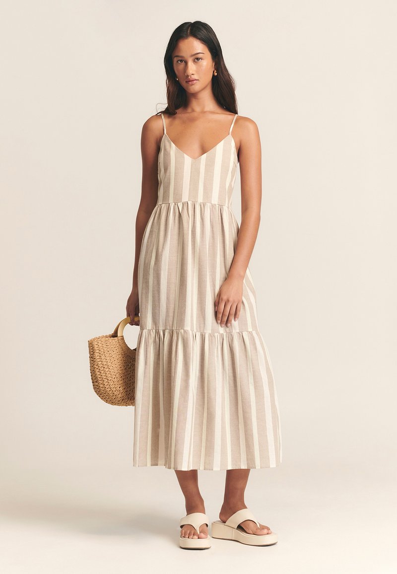 Abito in lino a righe con scollo a V, spalline sottili regolabili, orlo a balze, tonalità beige neutre, abbinato a una borsa in paglia intrecciata e sandali con zeppa.