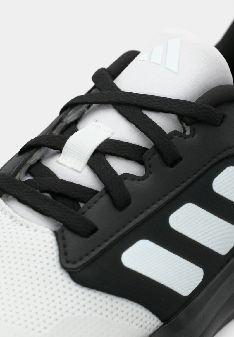 Zapatilla deportiva en blanco y negro con parte superior de malla, superposiciones texturizadas, cordones negros planos y tres rayas blancas en el costado. Destaca un lazo para los cordones.