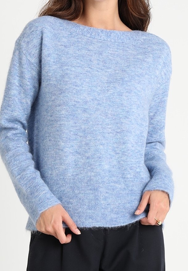 Femme portant un pull en maille duveteuse bleu clair à manches longues et un pantalon noir, les mains reposant près de l'ourlet du pull.