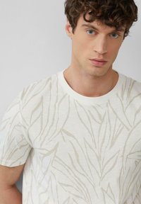 Korte mouwen, crew-neck t-shirt in lichtbeige met een subtiele, structuurrijke bladdessin op een witte achtergrond. Zachte stof en een ontspannen pasvorm.