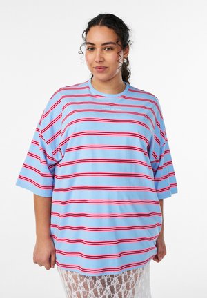 Kvinde med krøllet hår iført en løs blå skjorte med røde striber og hvid tekst "STATE OF EASE" stående foran en ensfarvet baggrund.