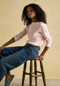 Mujer con cabello rizado que lleva una camiseta de manga larga a rayas rosas y blancas y jeans azules, sentada de lado en un taburete de madera.