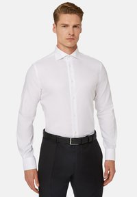 Camicia bianca a maniche lunghe con colletto classico, chiusura a bottoni e taglio elegante. Indossata con pantaloni neri e una cintura nera. Tessuto liscio.