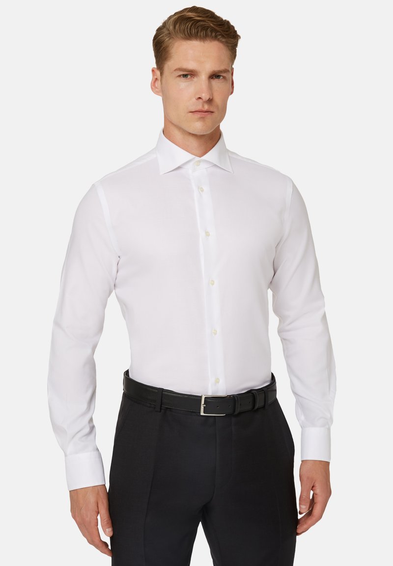 Camicia bianca a maniche lunghe con colletto classico, chiusura a bottoni e taglio elegante. Indossata con pantaloni neri e una cintura nera. Tessuto liscio.