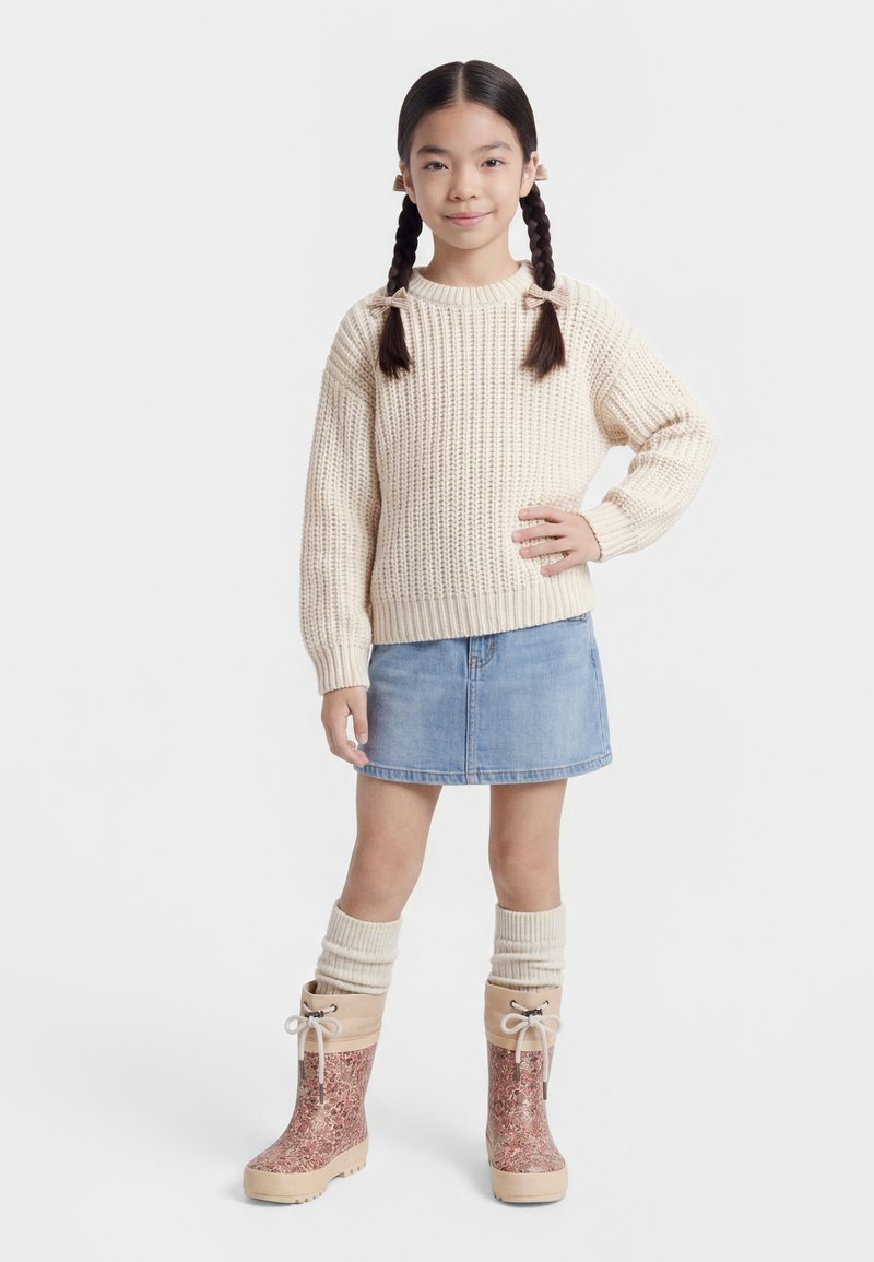 Mladé dievča s vrkočmi, oblečené v krémovom pletenom svetri, rifľovej sukni, krémových návlekoch na nohy a ružových členkových čižmách s potlačou, stojace s rukou na bedre.