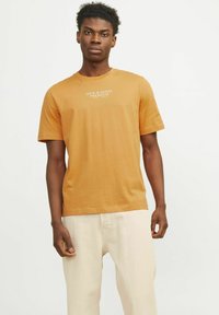 Polo majica s kratkimi rokavi v oranžni bombažni tkanini, ki vsebuje belo logotipno besedilo na prsih. V kombinaciji z svetlo bež hlačami.