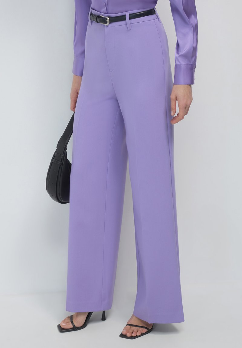 Lavendelweite Hosen aus glattem Stoff, mit hoher Taille und einem schwarzen Gürtel. Kombiniert mit schwarzen Riemchenschuhen und einer kleinen Handtasche.