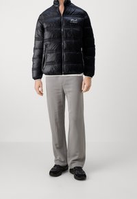 Sort pufferjakke med lynlås foran, vandret quiltet design og logo på brystet. Båret sammen med lysegrå bukser og sorte sneakers.