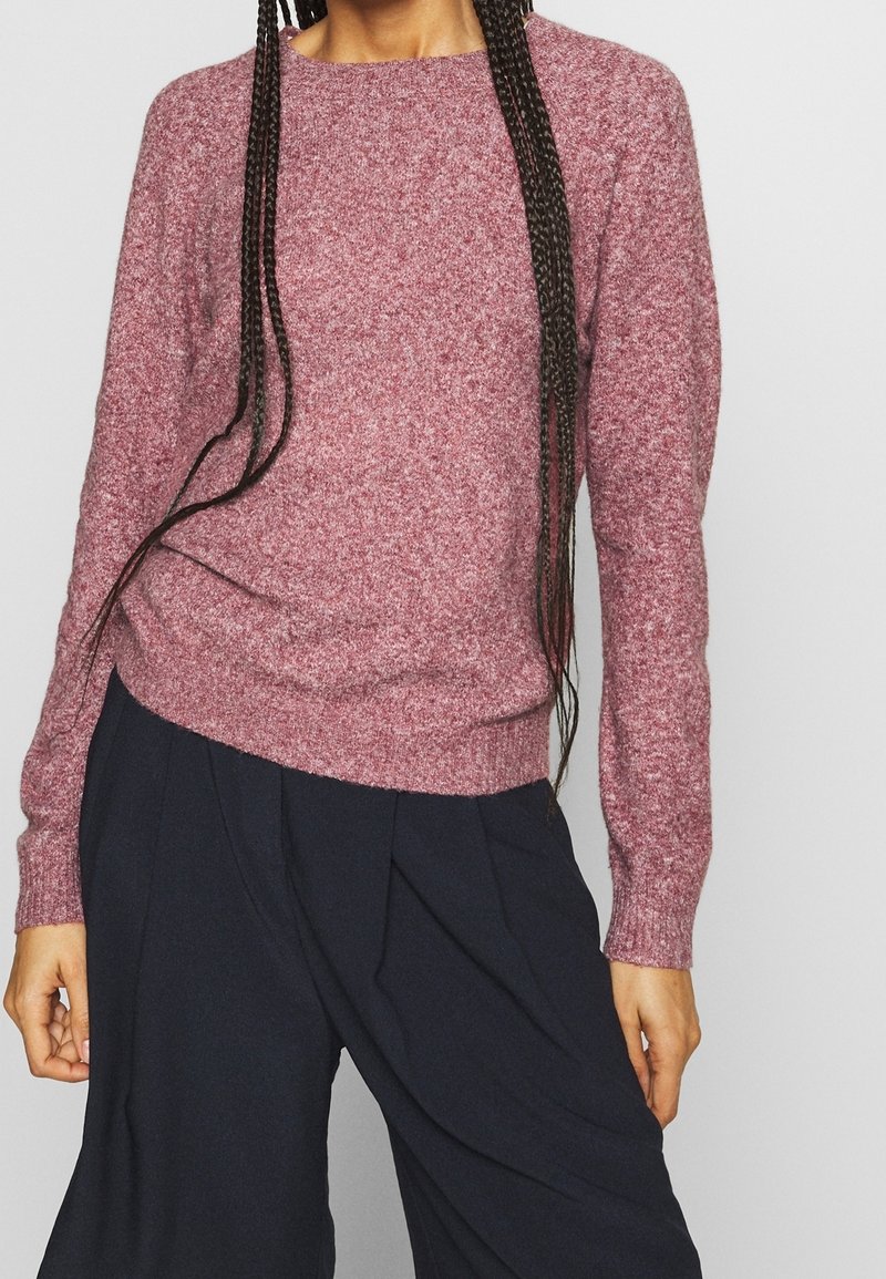 Personne portant un pull en maille rose poussiéreux à manches longues et un pantalon noir ample, avec de longs cheveux tressés sur les épaules.