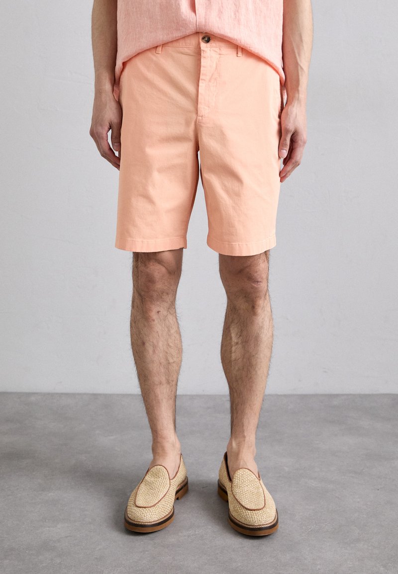 Pantaloni corti di cotone rosa chiaro con una vestibilità dritta e chiusura con bottone, abbinati a scarpe slip-on color tan con una texture intrecciata e dettagli marroni.