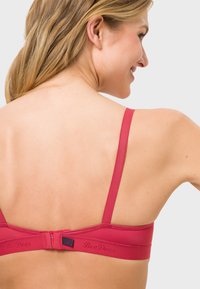 Red bra with adjustable straps, smooth texture, and hook closure. Features embossed logo along the band. Minimalistic design.