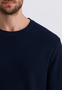 Marineblauwe gebreide trui met een textiele stof, ronde hals en geribde manchetten. Close-up opname met focus op het bovenlichaam.