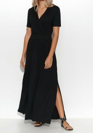 Mujer con vestido largo negro de manga corta con abertura lateral, combinado con sandalias planas negras con tachuelas, de pie sobre un fondo blanco.