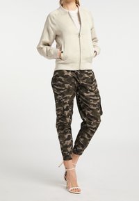Chaqueta bomber beige con cremallera oculta, puños acanalados y bolsillos laterales, combinada con pantalones cargo de camuflaje con puños elásticos y múltiples bolsillos.