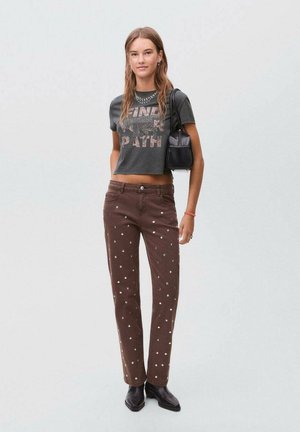 Pantalons en velours côtelé marron avec des ornements circulaires blancs, associés à un crop t-shirt graphique foncé. Le mannequin tient un sac à main noir.