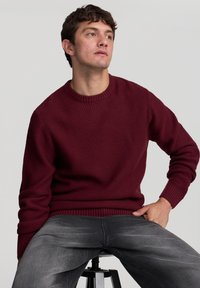 Maglione borgogna lavorato a maglia con scollo rotondo e polsini a costine, abbinato a jeans grigi. Il tessuto ha un aspetto testurizzato e intrecciato.