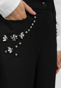 Pantalons noirs avec des embellissements en cristal sur la poche avant, d'une texture lisse et d'un design ajusté. Les cristaux varient en forme et en taille.