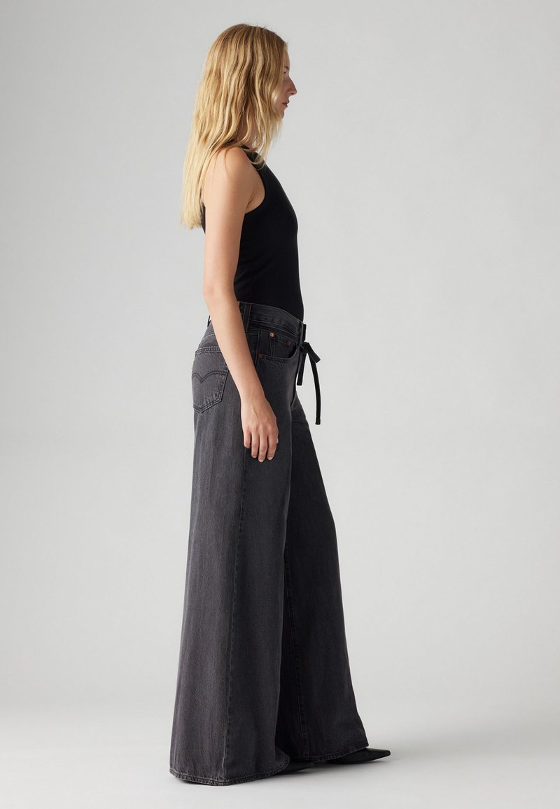 Schwarz-mit einem hohen Bund ausgestellte Damenjeans mit Frontbindedetail und Taschen. Das Model trägt ein schwarzes ärmelloses Top und zeigt ein Seitenprofil.